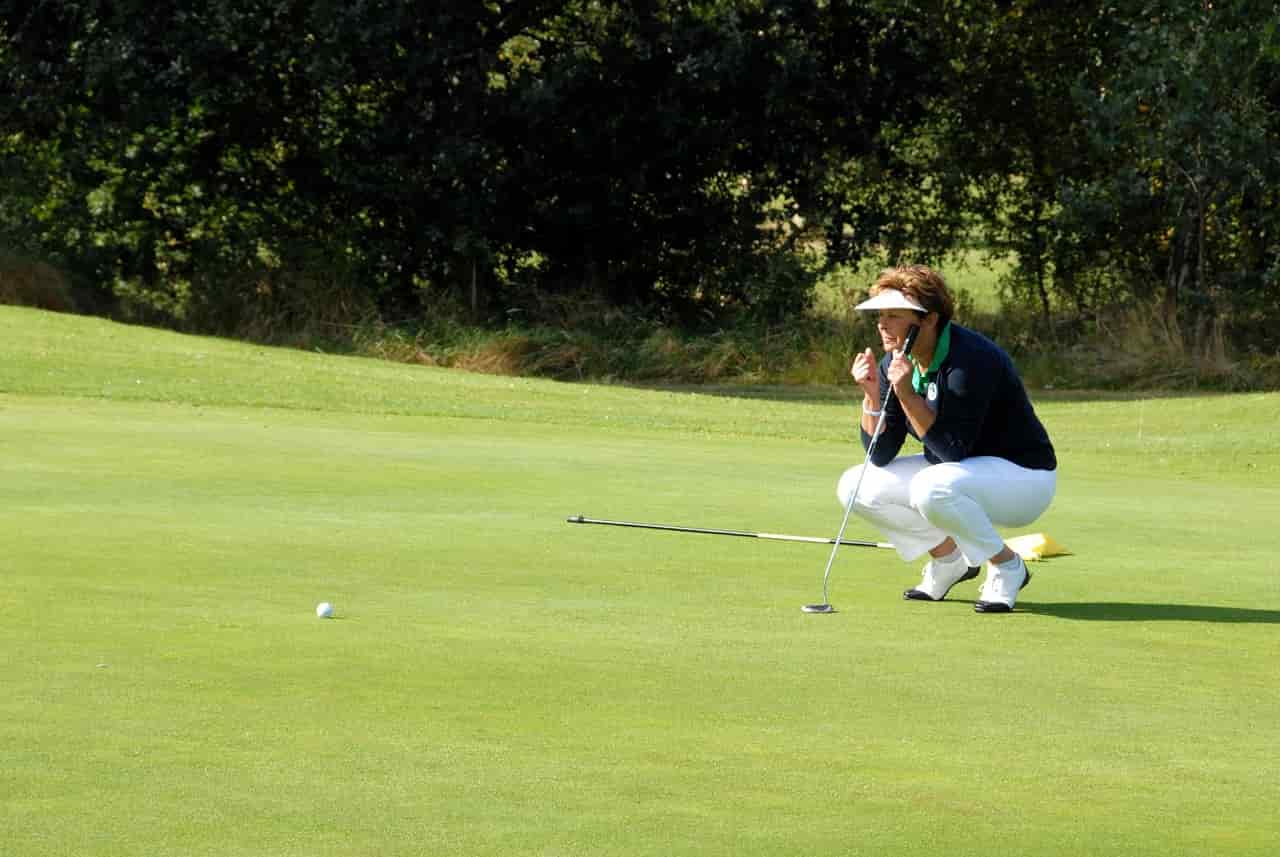 Frau hält auf dem Golfplatz bei einem Kurzurlaub in Oberbayern einen Golfschläger in der Hand und kniet auf grünen Rasen.
