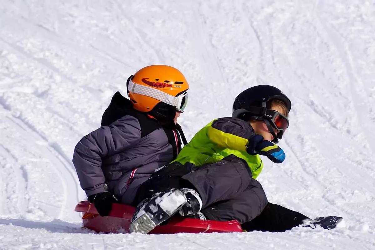 Zwei Kinder in Winterkleidung und mit Helm auf einem Rodel im Schnee in Oberbayern.