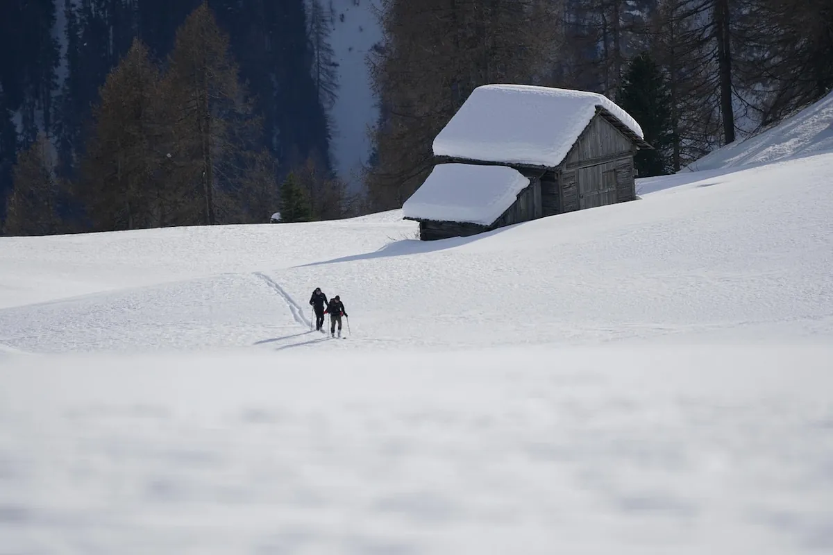 zwei skitourengeher beim aufstieg im pulverschnee. im hintergrund eine verschneite berghütte in oberbayern