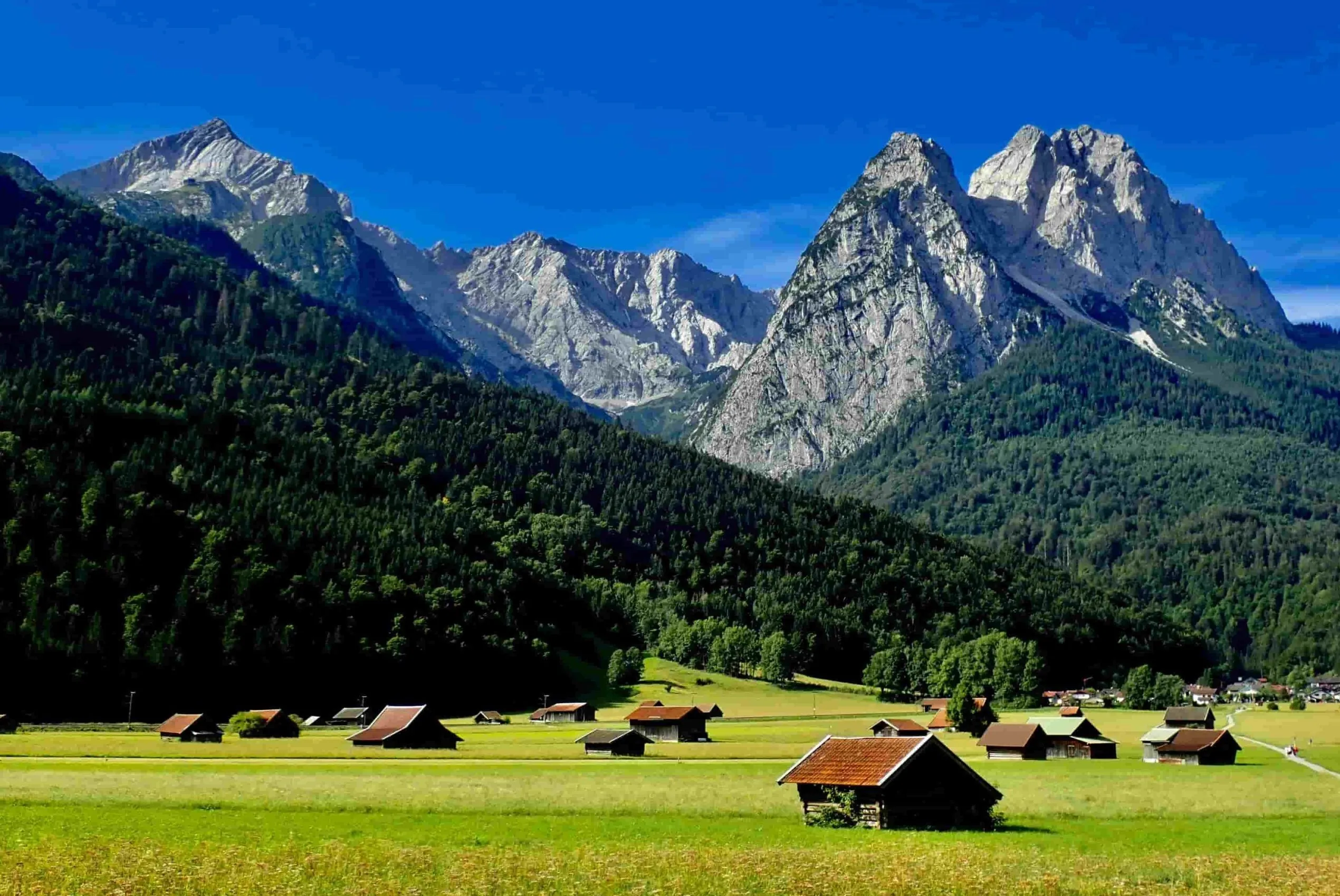 Grüne Wiese mit kleinen Holzhütten in der Zugspitzregion. Im Hintergrund Bergkulisse Zugspitze und Alpspitze.